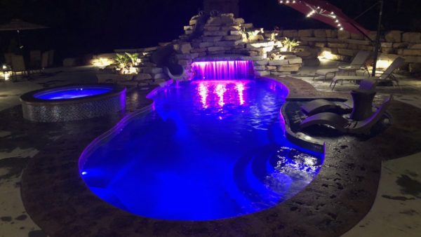 Swimming pool with integrated spa at night glowing with purple light. Pool lounge chairs on the side.