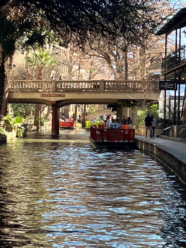 Tour boats travel along a tree-lined urban canal beneath a stone pedestrian bridge, with people walking along the riverside path and sunlight filtering through the branches overhead.